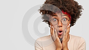 Photo of stupefied emotional black woman with bugged eyes, keeps hands on cheeks, stares through spectacles, models