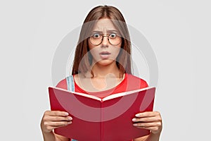 Photo of stupefied emotional young Caucasian woman looks with stupefaction at camera, holds red book, has to learn much