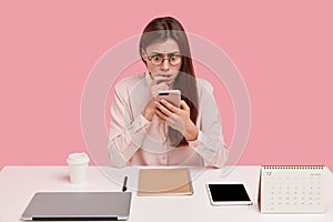 Photo of stupefied European woman browses internet on modern cell phone, has scared expression, poses at workspace