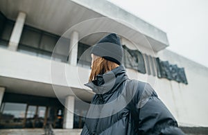 Photo of a stylish woman in a warm winter jacket and hat standing on the street against the backdrop of architecture and looking