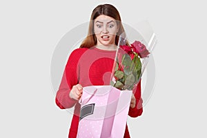 Photo of surprised terrified young woman has stupefied facial expression while opens gift bag, holds roses, dressed in red clothes