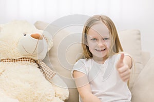 Photo of teddy bear and little girl showing thumb up at the camera.