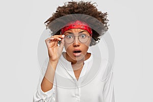 Photo of terrified beautiful woman keeps hand on rim of spectacles, stares with scrupulous look at camera, dressed in photo