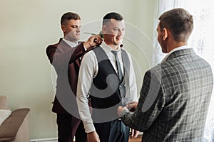 Photo of three men in classic suits. A handsome young man mends the shirt of another man, standing behind him. Business style.