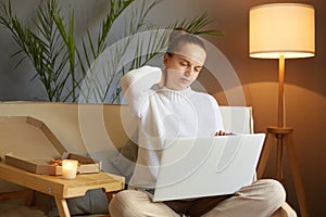 Photo of tired exhausted young adult woman wearing white sweater sitting on sofa and working on laptop in home interior, having