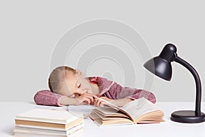 Photo of tired small kid takes nap on desktop, wants sleep, has to make homework, sleeps near opened book, holds pencil for writin