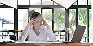 Photo of a tired woman keeps a hand on her head at the wooden working desk after finishing work hard.