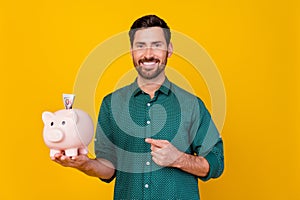 Photo of toothy beaming man dressed dotted shirt indicating at money piggy box with banknote inside isolated on yellow