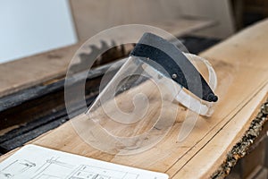 Photo of transparent protective face mask lying on the stack of lumber with blurred table saw on background