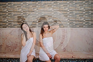 Photo of two beautiful women wearing white towels relaxing in hamam