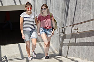 Photo of two female lesbians hold hands, have walk in city, stand near subway, wears casual t shirts and jean shorts, have happy e
