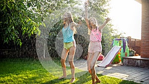 Image of two happy laughing teenage girls jumping and dancing under warm summer rain at house backyard garden. Family