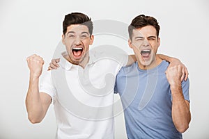 Photo of two happy mates 30s wearing casual t-shirt and jeans hugging and screaming while clenching fists in joy, isolated over w