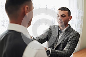 Photo of two men in classic suits. A handsome young man mends the shirt of another man standing in front of him. Business style.