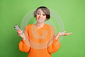 Photo of uncertain young woman shrug shoulders hold smartphone blogger isolated on green color background