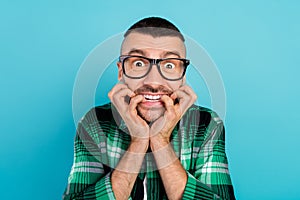 Photo of unhappy upset scared shocked young man bite fingers wear glasses horrified isolated on blue color background