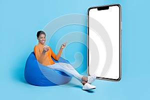 Photo of unisex person sit bean bag hold phone direct poster empty space wear yellow shirt isolated blue color photo