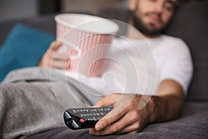 Photo of unshaved man holding popcorn bucket while sleeping on couch in living room