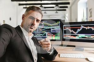 Photo of unshaved man taking selfie while working in office on computer with graphics and charts at screen