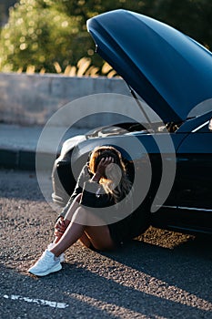 Photo of upset woman sitting on asphalt next to broken car with open hood