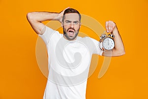 Photo of uptight man 30s in casual wear holding alarm clock, isolated over yellow background