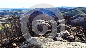 A photo of vegetation being harmed by fire damage, mountain rocks, burnt bushes and a mountain range in the distance