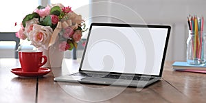 Photo of white blank screen computer laptop putting on working desk with bunch of flowers, notebook and pencil holder.