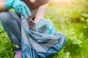Photo of woman in rubber gloves picking up dirty plastic bottle in bag on green lawn