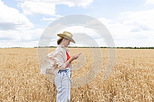 Photo of woman in straw hat looking on cellphone while walking on wheat field at summer day