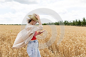 Photo of woman in straw hat looking on cellphone while walking on wheat field at summer day