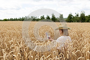 Photo of woman in straw hat looking on cellphone while walking on wheat field at summer day