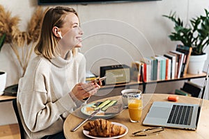 Photo of woman using cellphone and earbuds while having breakfast at home