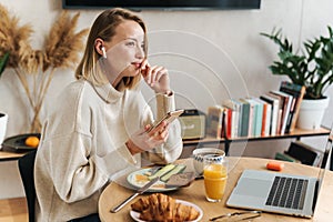 Photo of woman using cellphone and earpods while having breakfast