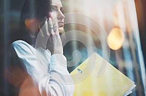 Photo woman wearing white shirt, talking smartphone and holding business files in hands. Open space loft office. Panoramic windows