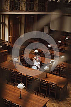 Photo of a woman in white clothes sitting alone in a public library at a table with a book on her head. Vertical