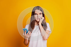 Photo of worried lady bite nails hold phone wear white t-shirt posing on yellow background