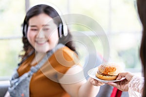 Photo of write hand holding a white plate of hamburger with soft focus on smiling fat woman with headphones use the left hand to