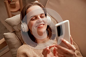 Photo of young adult Caucasian woman wearing headphones holding cell phone while lying on sofa in her apartment, using mobile