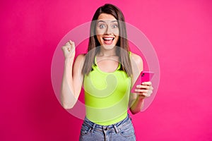 Photo of young amazed lady hold cell phone open mouth raise fist wear green top isolated vibrant pink color background