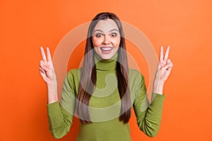 Photo of young attractive smiling excited happy girl showing v-sign wear green turtleneck isolated on orange color