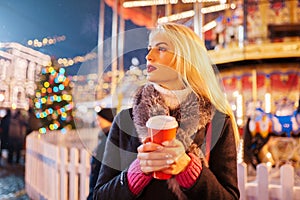 Photo of young blonde with glass in hands on background of carousel in evening for walk