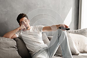 Photo of young bored man sitting on couch at home, and changing