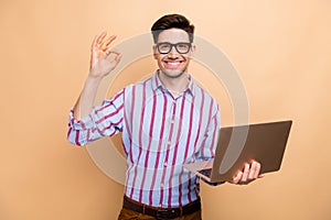 Photo of young cheerful business guy manager show okey gesture got deal in ecommerce using laptop isolated on beige