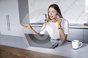 Photo of a young cheerful happy woman at home at the kitchen using laptop computer talking by mobile phone