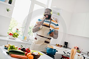 Photo of young excited guy have fun enjoy cooking listen radio headphones singer mic spatula sound cook cuisine indoors