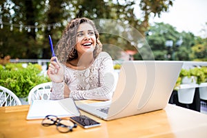 Photo of young excited latin curly woman indoors using laptop computer writing notes Looking aside have an idea in cafe