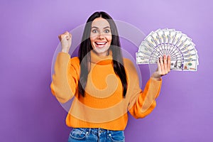 Photo of young funky overjoyed woman brunette hair wear orange sweater fist up hold much money win lottery isolated on