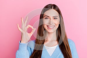 Photo of young good mood positive girl in blue cardigan showing okey gesture smiling on camera isolated on pink color