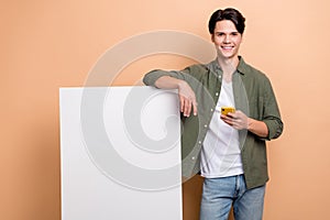 Photo of young handsome positive guy wearing trendy khaki shirt with jeans hold phone recommend banner new app isolated