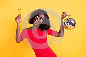 Photo of young happy smiling african girl in sunglasses hold disco ball drink champagne isolated on yellow color